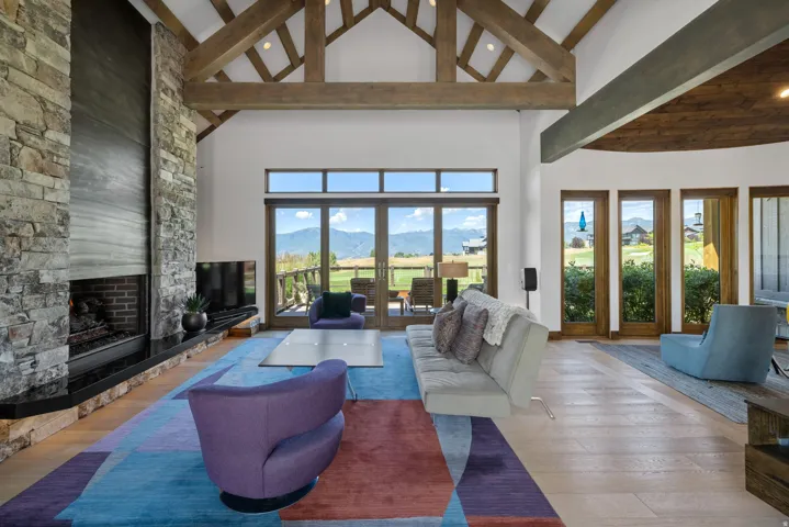 Spacious living area featuring a stone fireplace with a black hearth, vaulted ceilings with exposed wood beams, and light wood-finish flooring