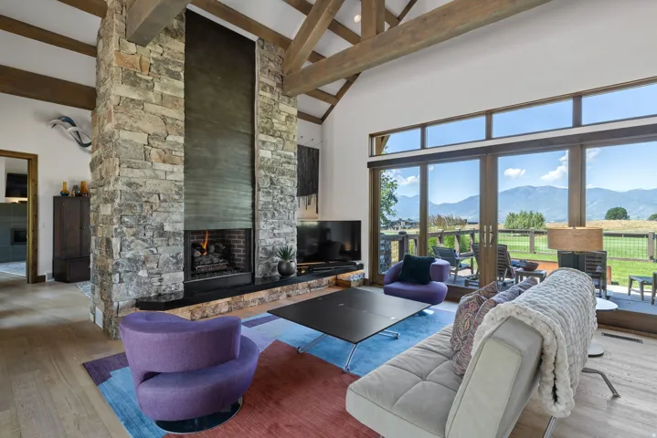 Vaulted ceiling with exposed wood beams, a stone fireplace with a dark metal surround, and an expansive wall of glass doors opening to a patio