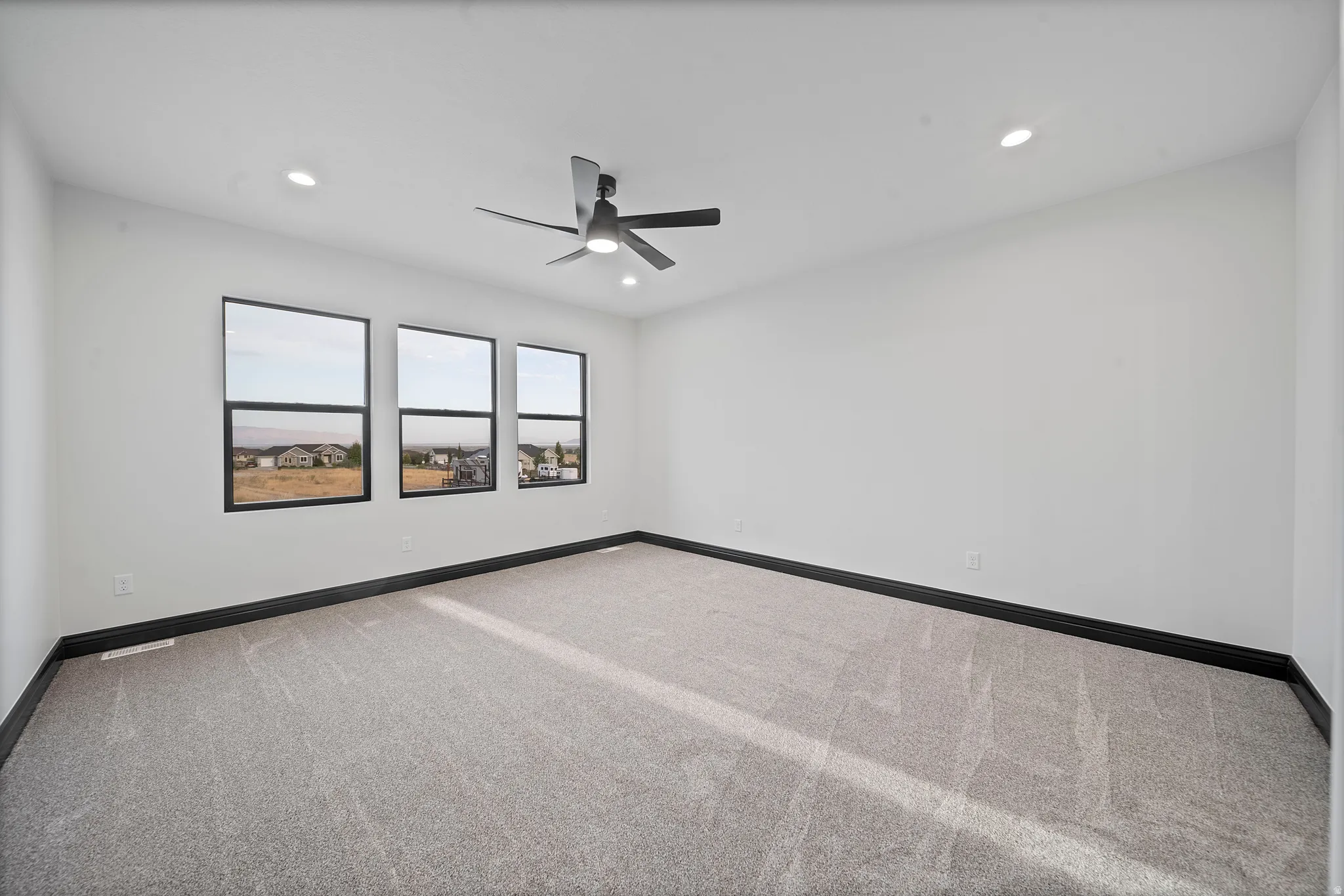Spacious room with light gray carpeting, three large windows with black frames, a modern ceiling fan, and recessed lighting