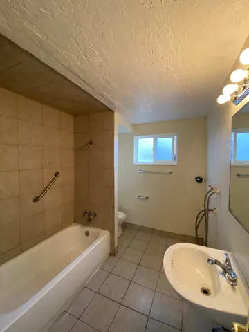 Bathroom featuring a pedestal sink, tiled flooring, bathtub with tiled surround, grab bar, and a frosted window