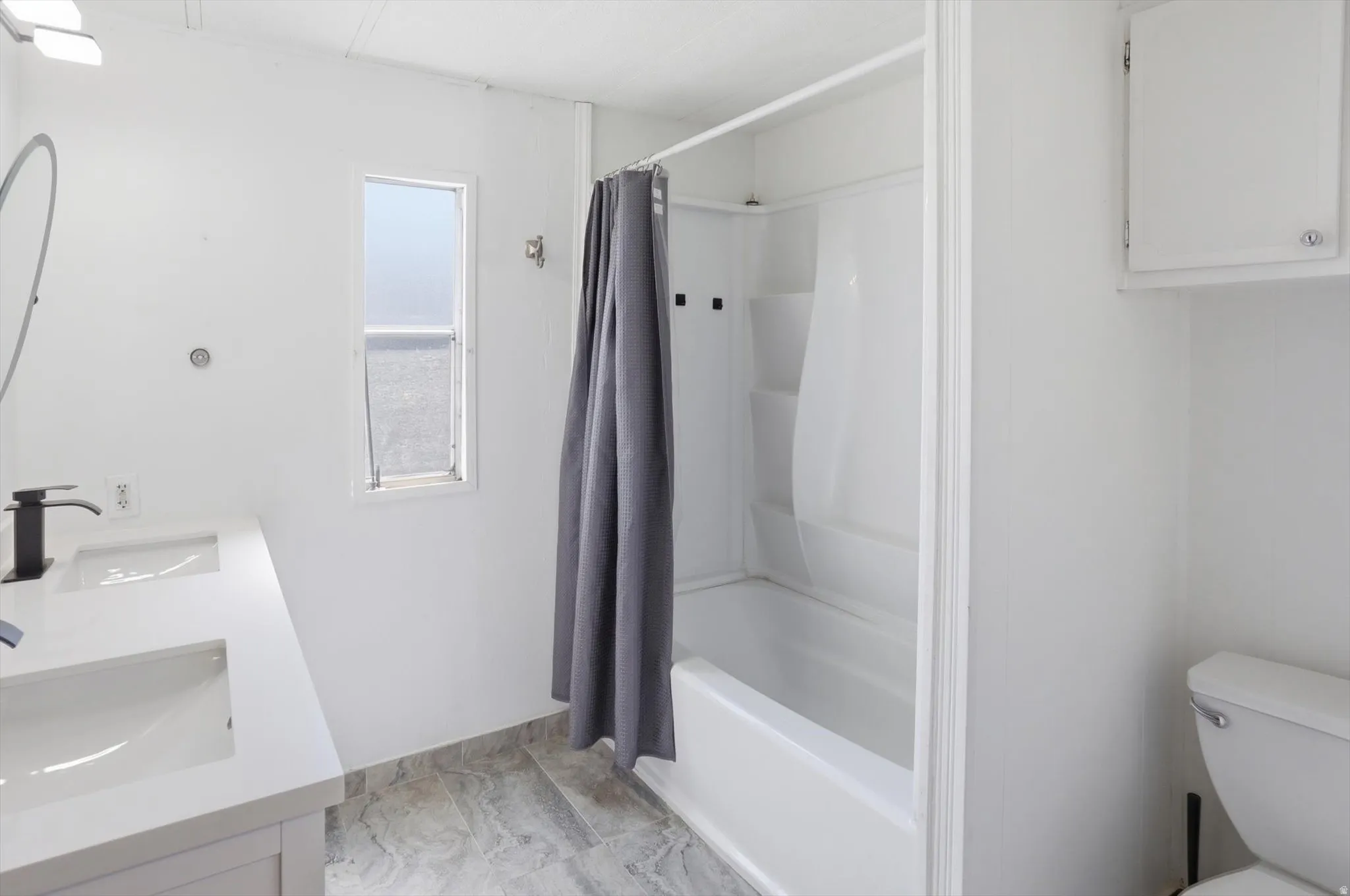 Bathroom featuring a dual-sink vanity with white countertops and matte black faucets, a white bathtub with an integrated shower, and light-colored tile flooring
