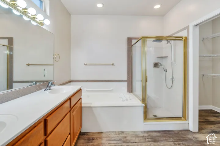 Bathroom featuring a double vanity with light wood cabinetry and integrated sinks, a built-in bathtub, a walk-in shower with a gold-tone frame, and wood-finish flooring