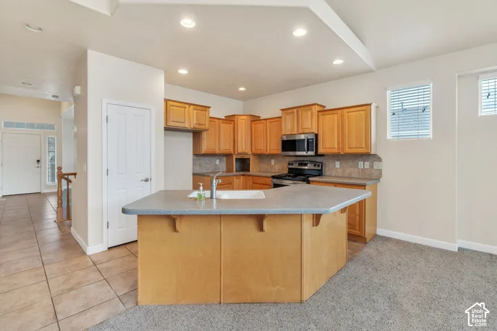 Open-concept kitchen featuring light wood cabinetry, a center island with sink, stainless steel appliances, recessed lighting, and a neutral tile backsplash