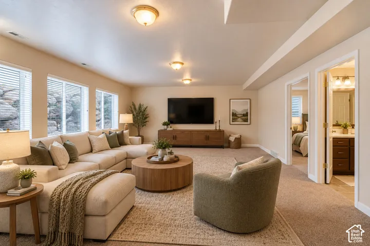 Spacious primary living area featuring a curved sectional sofa arrangement, wood-finish media console with integrated storage, recessed ceiling lighting, large multi-pane windows with plantation shutters, neutral carpet flooring, and direct access to adjo