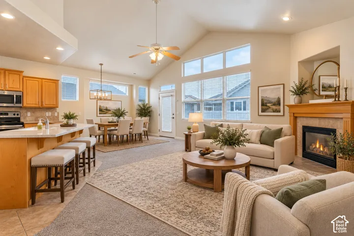Open-concept living area featuring a vaulted ceiling with a ceiling fan, a gas fireplace with a wood mantel, a kitchen island with seating, and a dining area with a chandelier