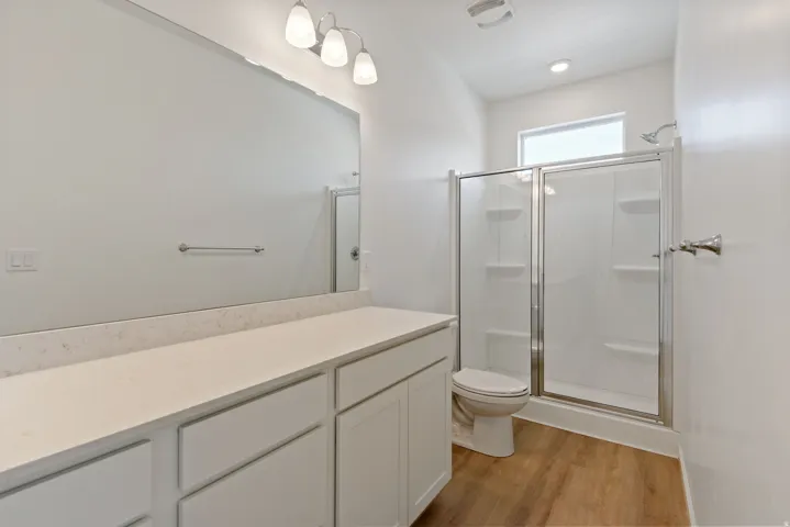 Bathroom featuring a wood-finish floor, a white vanity with a light-colored countertop, an expansive wall mirror, a glass-enclosed shower, and a white toilet