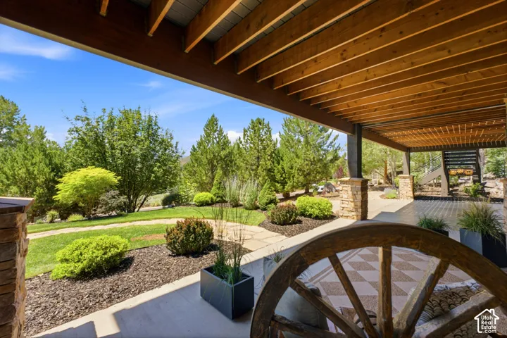 Covered patio featuring exposed wood beam ceiling, stone-wrapped columns, a concrete floor, and landscaping with mature trees