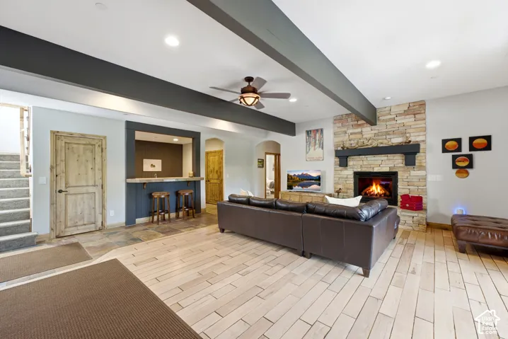 Spacious living area featuring a stone-clad fireplace with a dark wood mantel, a built-in wet bar, wood-finish flooring, a ceiling fan with integrated lighting, and exposed structural beams