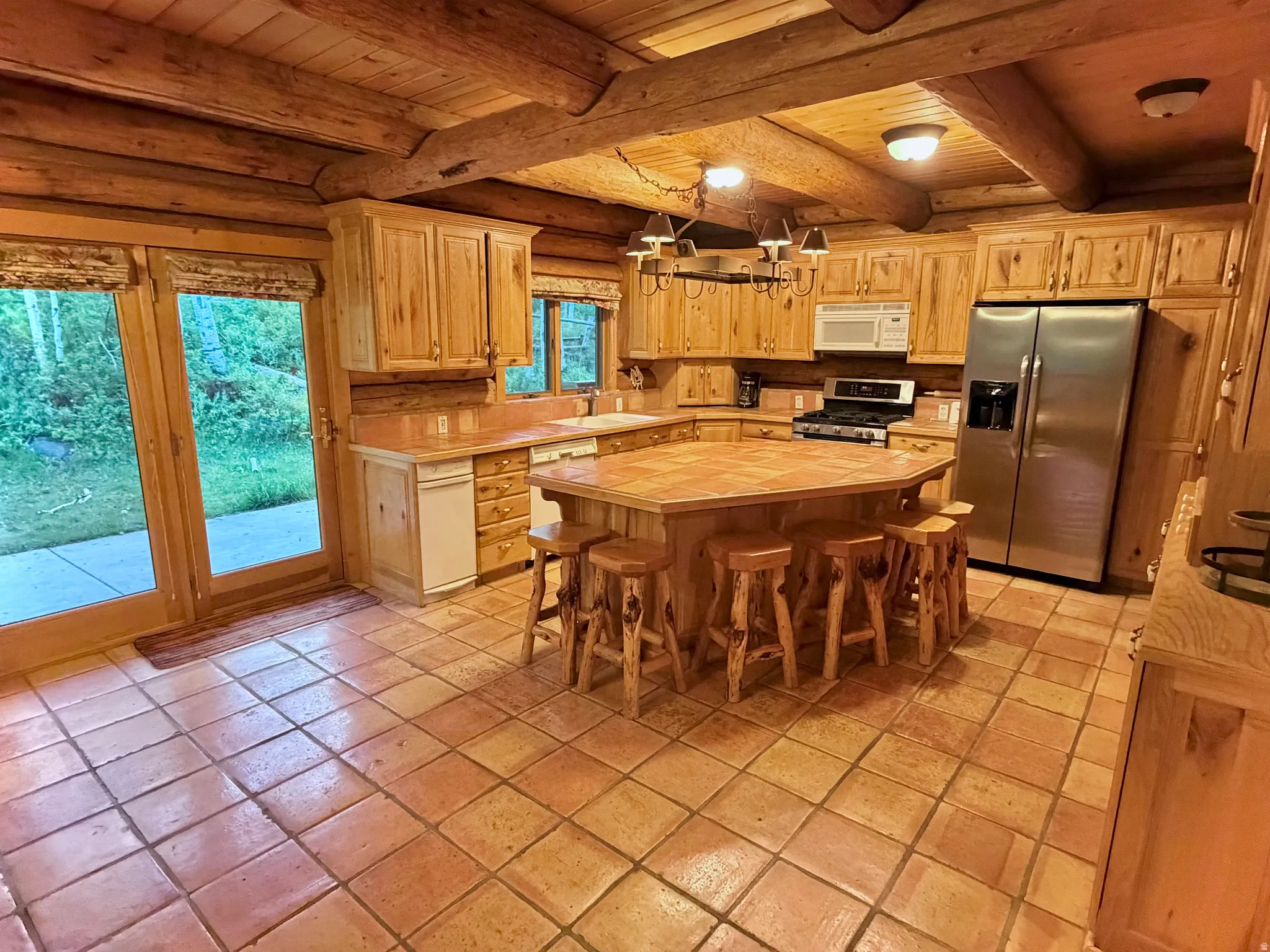 Kitchen featuring natural wood cabinetry, stainless steel appliances, a large center island, terra cotta tile flooring, and exposed log beam ceilings
