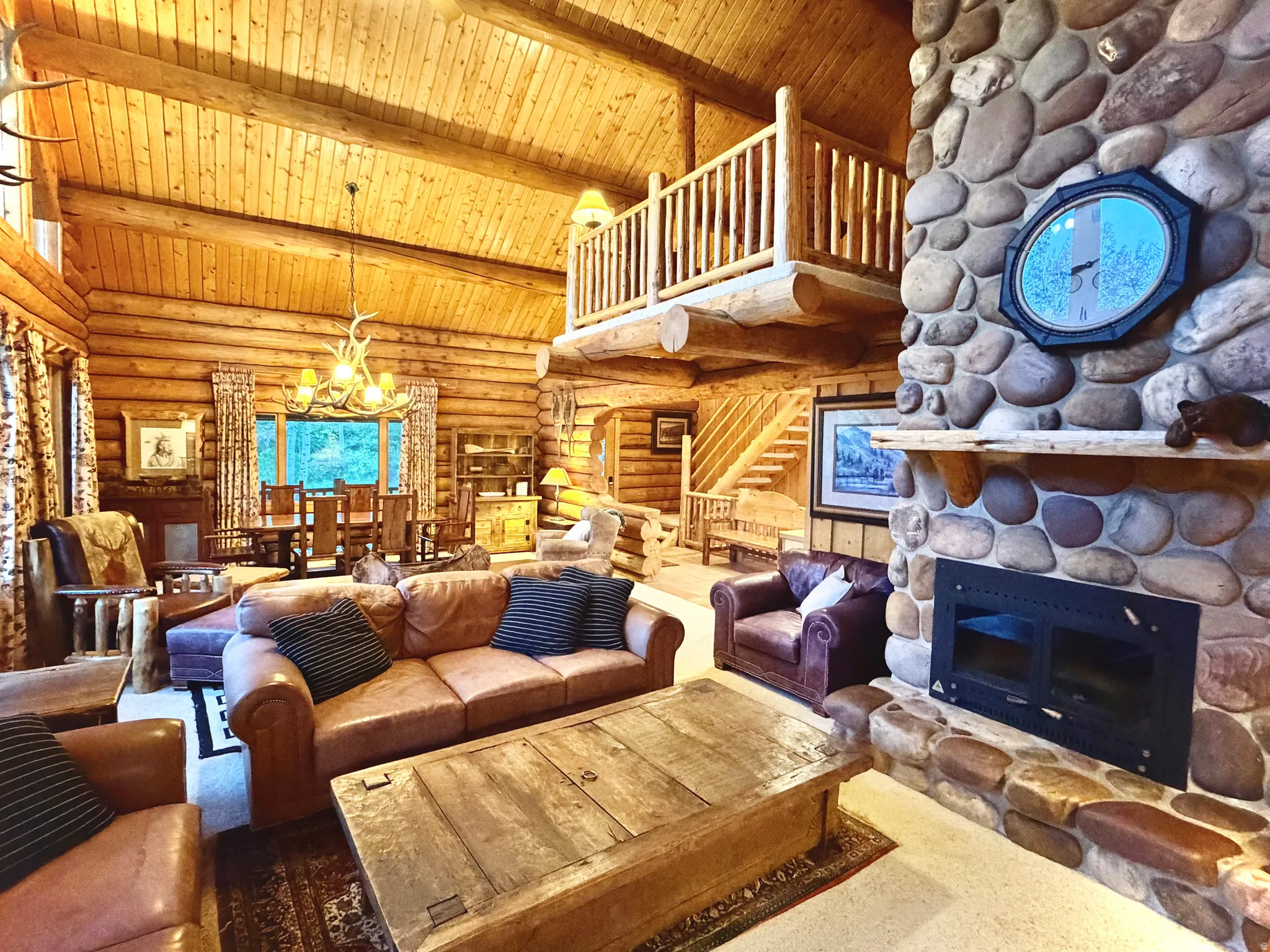 Expansive great room featuring a floor-to-ceiling river rock fireplace, exposed log walls, high vaulted ceilings with wood-plank finishing, and a prominent wood staircase leading to an upper-level balcony