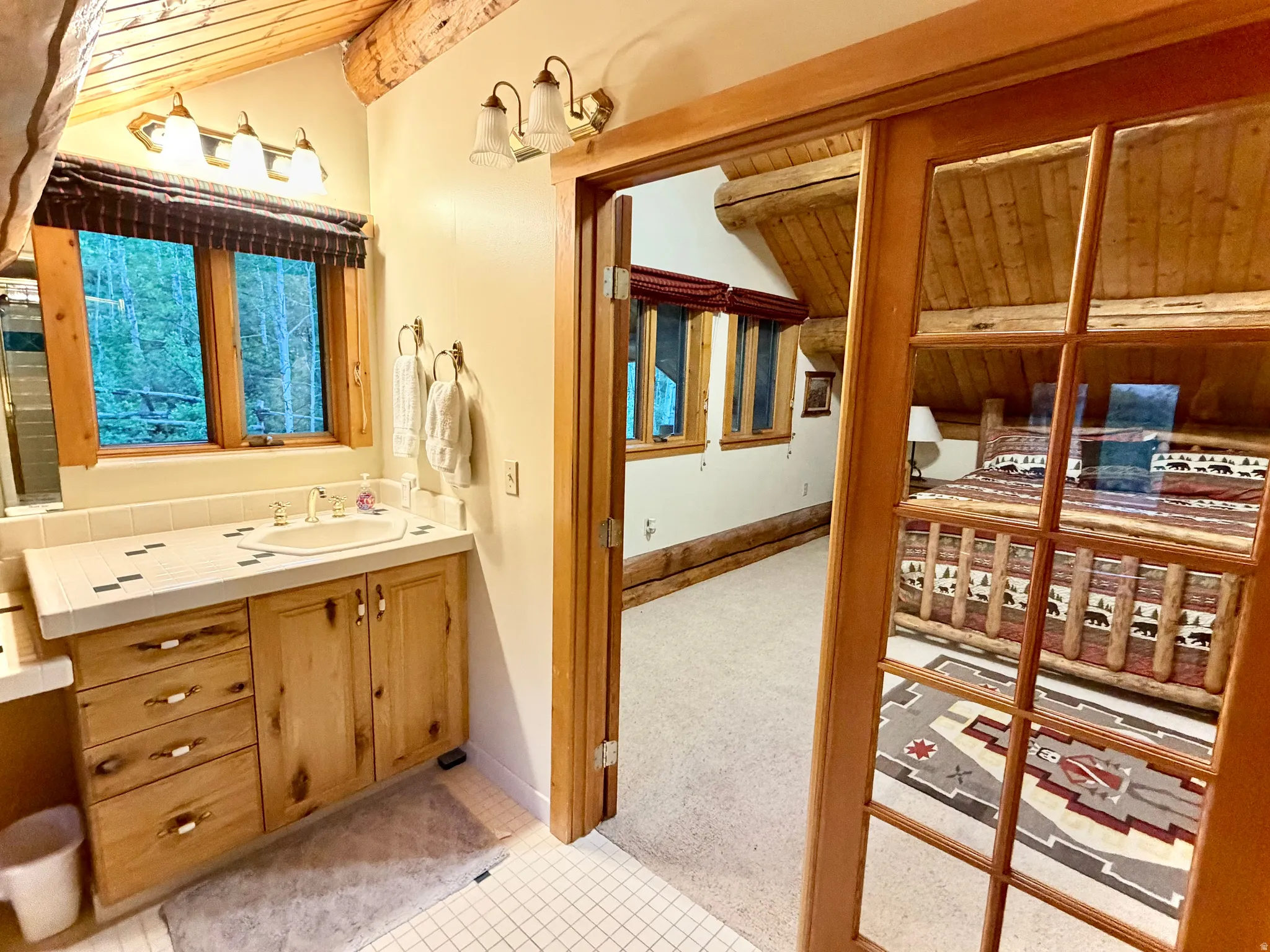 Bathroom vanity with a white tile countertop, integrated sink, light wood cabinetry, and a three-light fixture