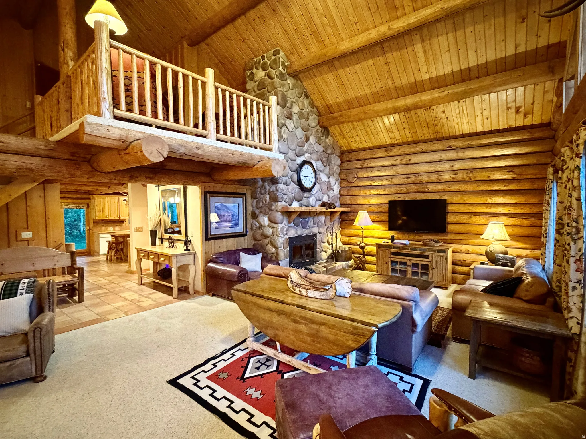 Open living area featuring vaulted wood-plank ceilings, log-beam construction, a stone fireplace with a wood mantle, and a visible loft space