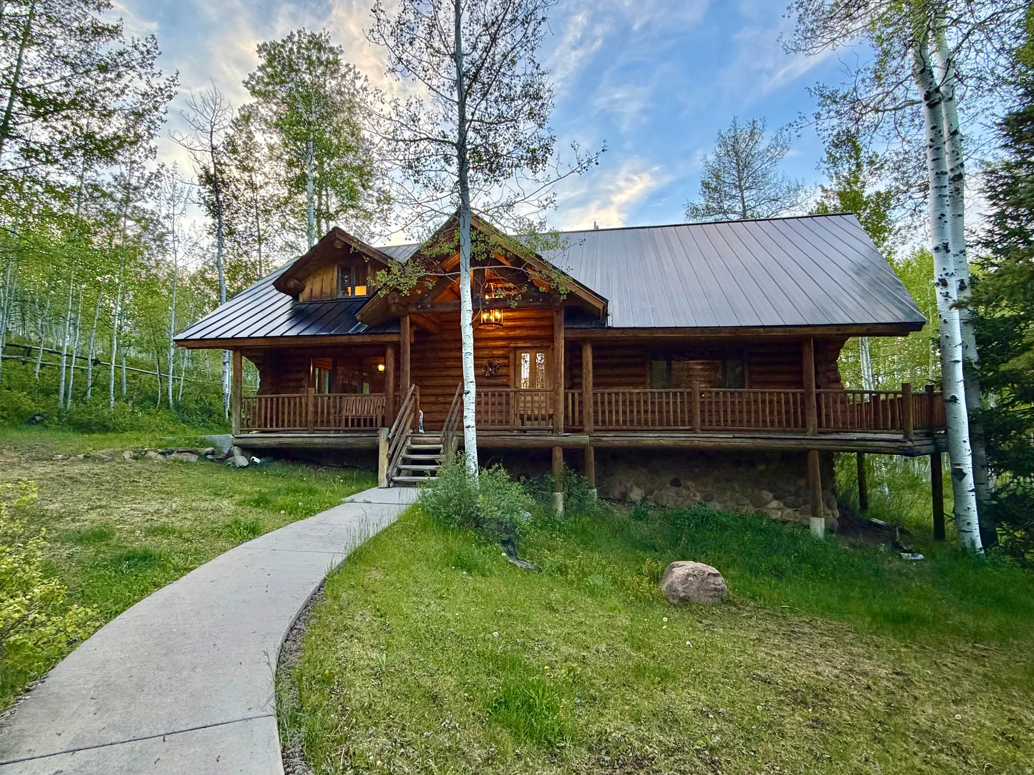 Log cabin exterior featuring a dark metal roof, wrap-around porch with wood railings, and a concrete pathway