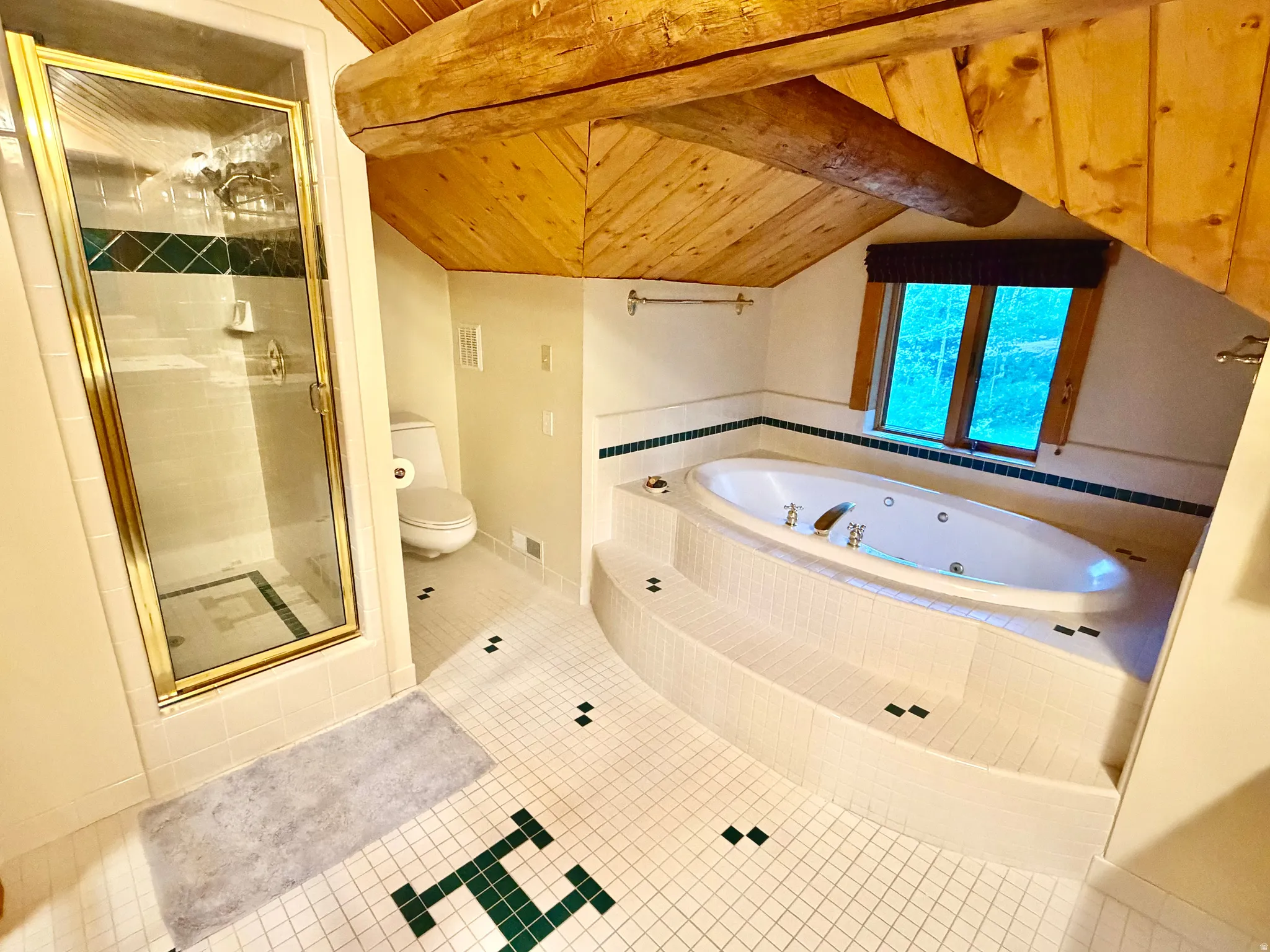 Bathroom featuring a jetted tub set into a tiled platform, a glass-enclosed shower with gold-tone fixtures, a toilet, a wood-plank ceiling with exposed log beams, and a window with a valance