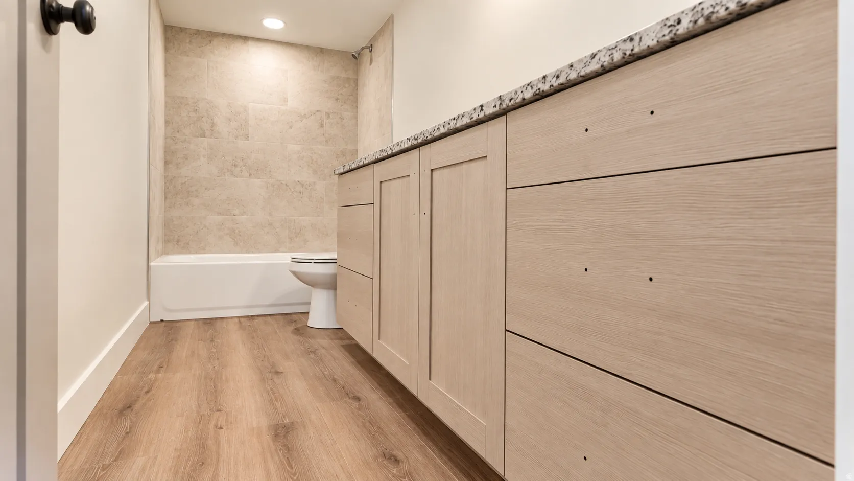 Wood-finish flooring, light-toned cabinetry, speckled countertop, tiled bathtub surround, and recessed lighting