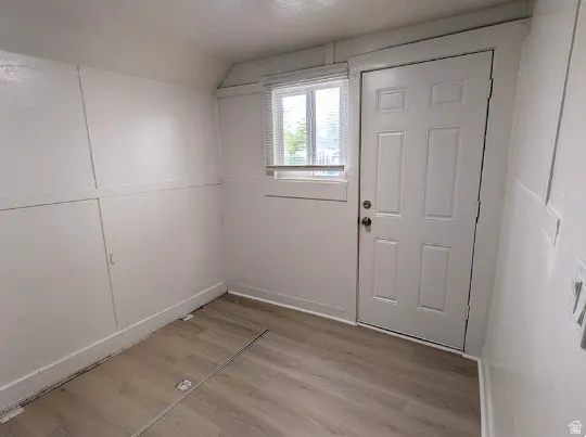 White paneled walls with wood-finish flooring, featuring a window with blinds and a paneled door