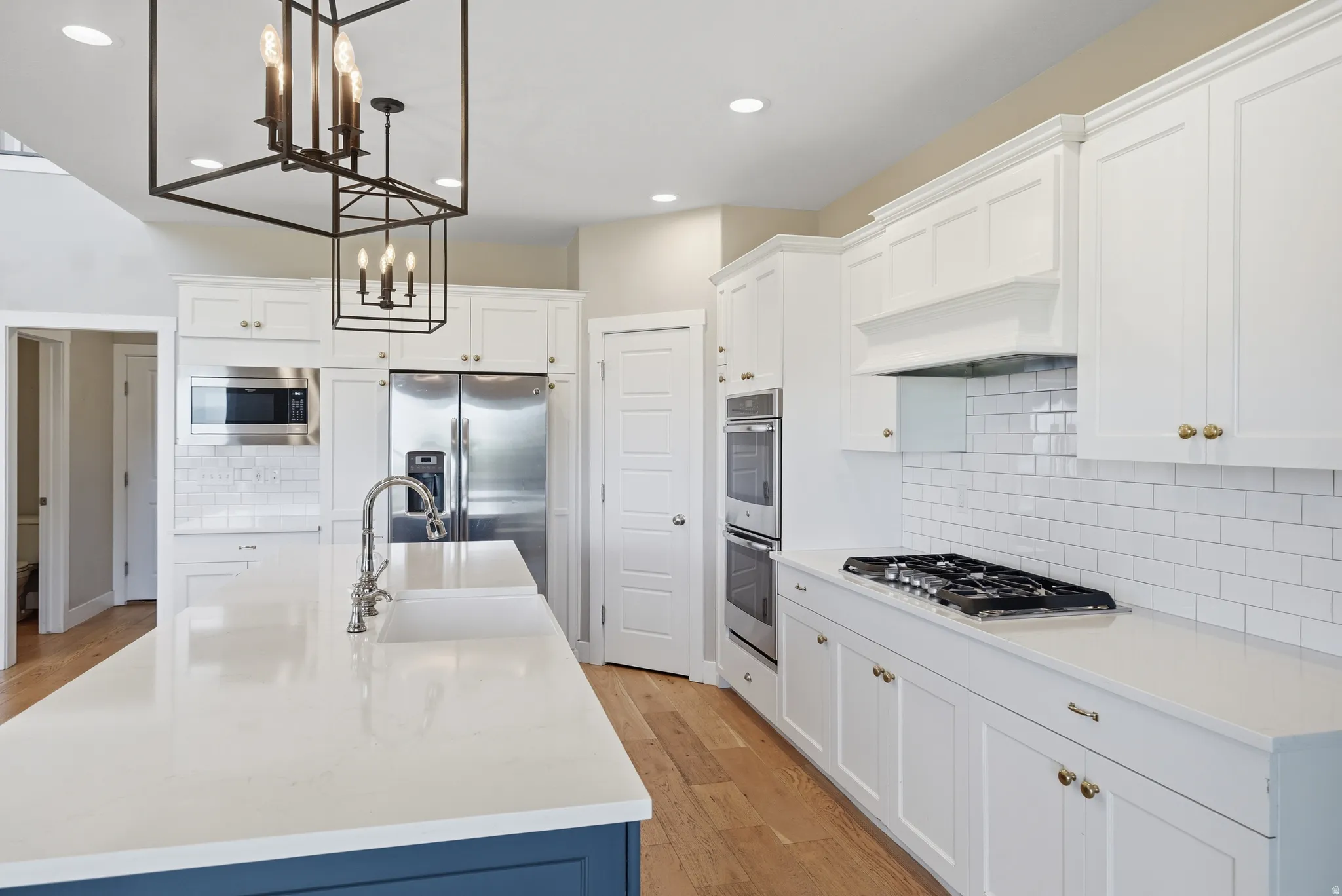 Kitchen featuring a large island with an integrated sink, stainless steel appliances, white shaker cabinetry, white subway tile backsplash, and wood-finish flooring