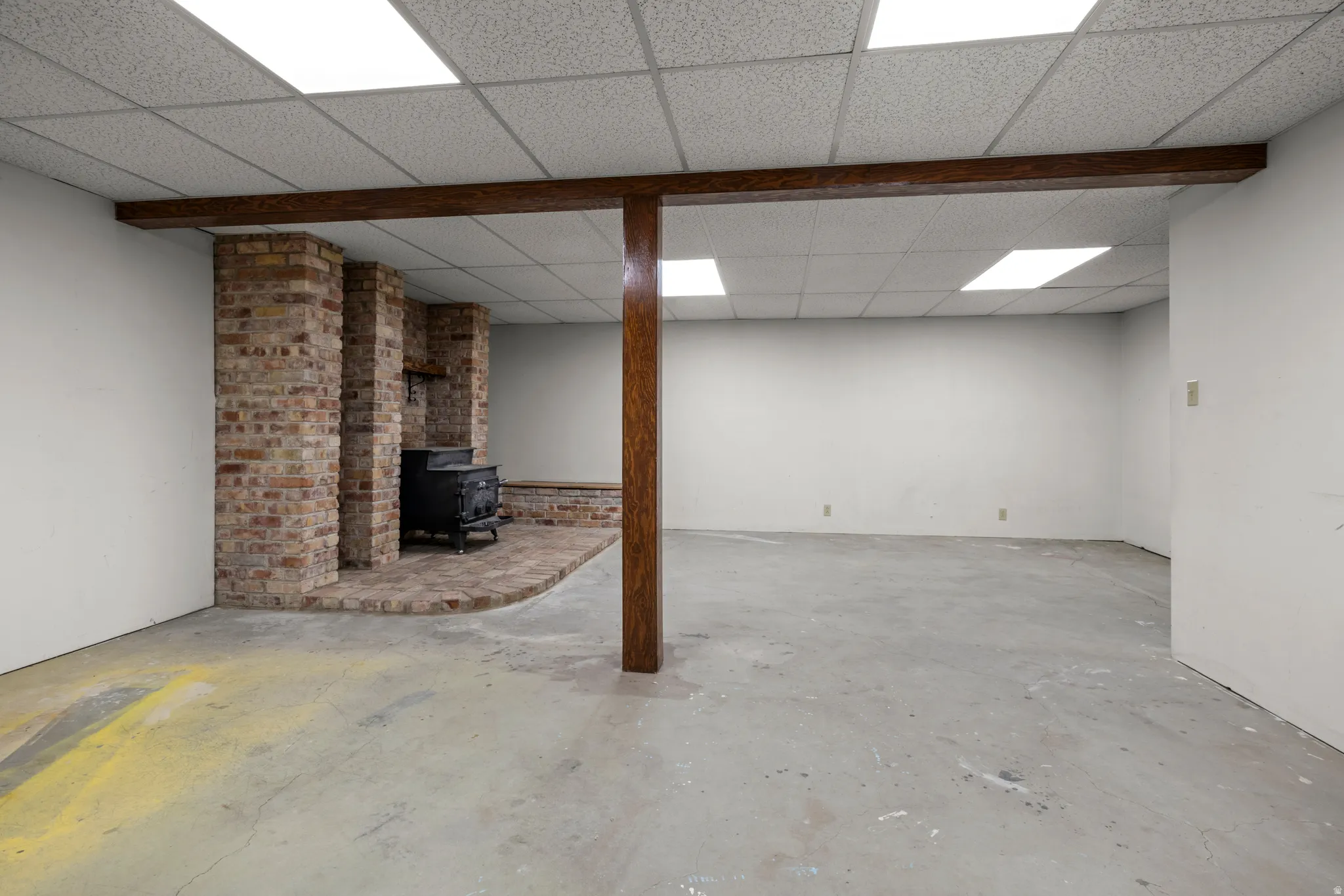 Spacious room featuring a brick-clad wood-burning stove with a matching brick hearth, exposed wood beam, and suspended acoustic tile ceiling