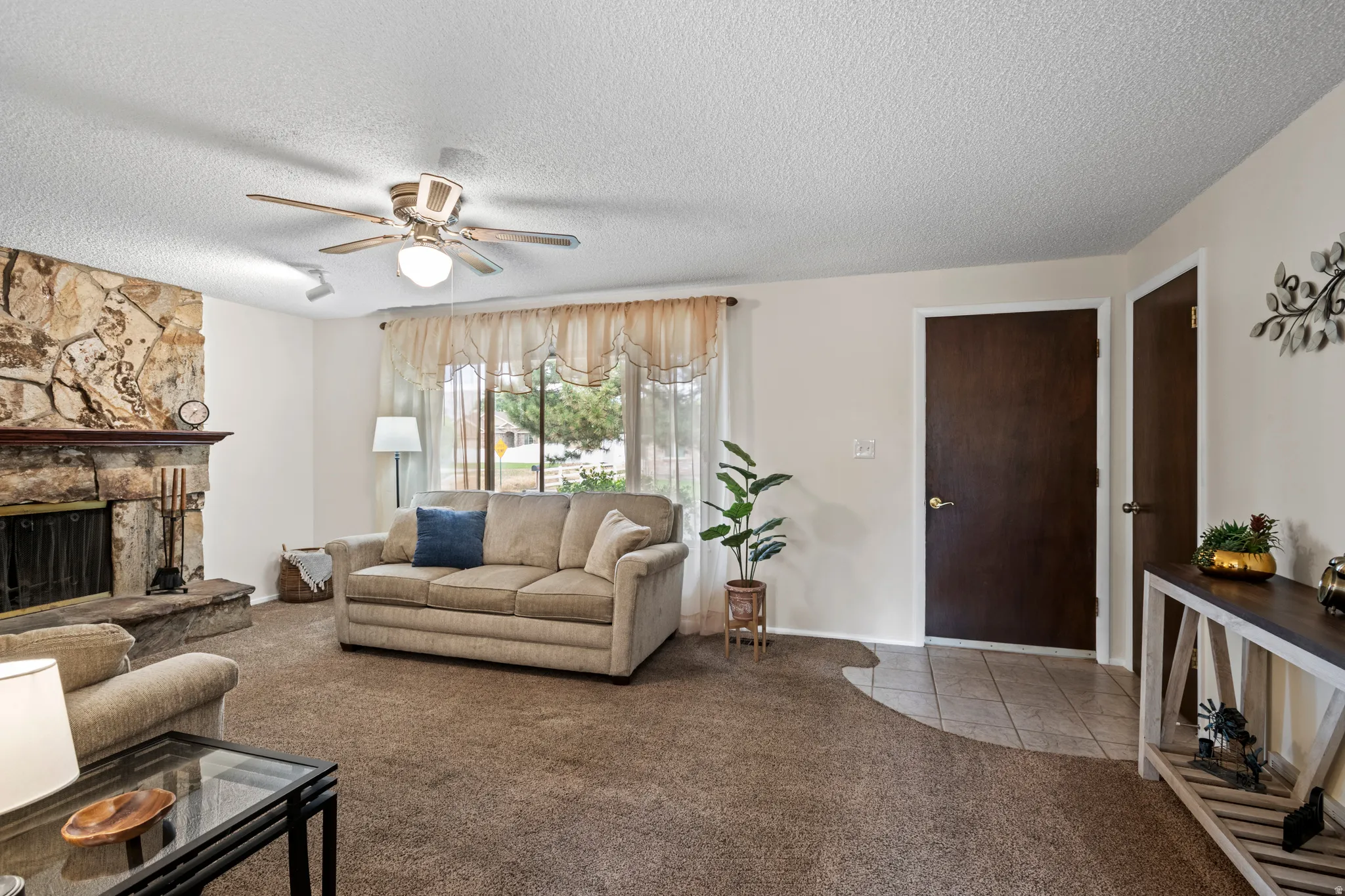Living area featuring a stone fireplace with a raised hearth, broad window with sheer treatments, and a ceiling fan