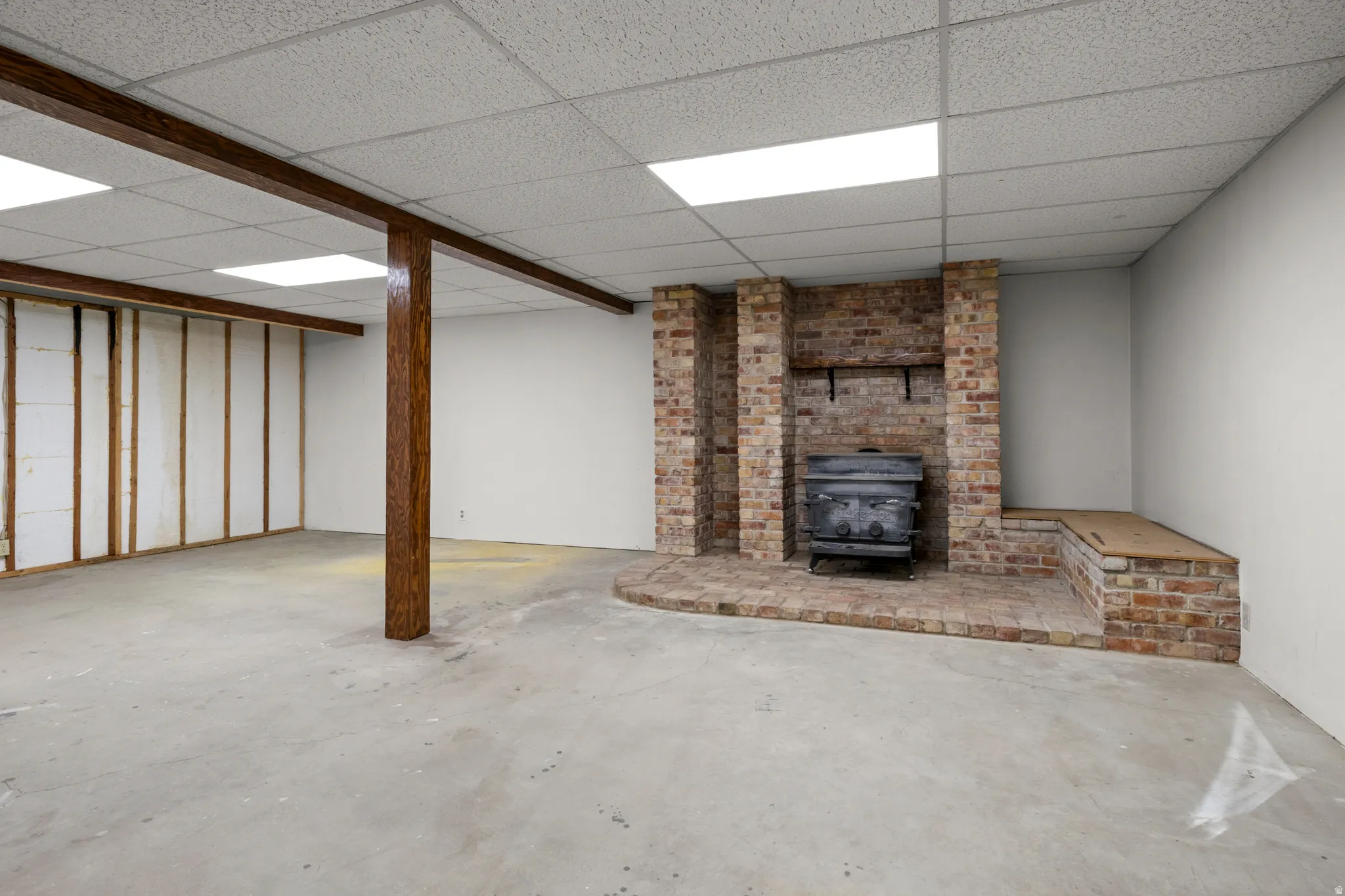 Spacious unfinished area featuring a brick-clad fireplace with a wood-burning stove insert, wood-finish support beams, and recessed lighting