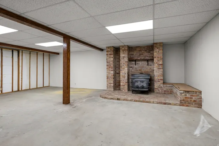 Spacious unfinished area featuring a brick-clad fireplace with a wood-burning stove insert, wood-finish support beams, and recessed lighting
