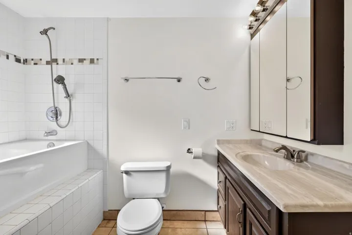 Bathroom featuring a bathtub with white tile surround, a built-in vanity with a light-toned countertop and dark wood-finish cabinetry, a chrome faucet, and a wall-mounted mirrored medicine cabinet