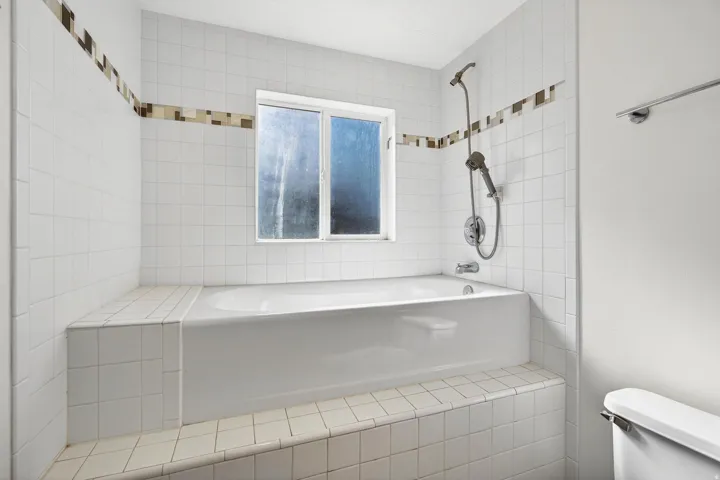 White tiled bathroom featuring a built-in bathtub with integrated showerhead and a window above