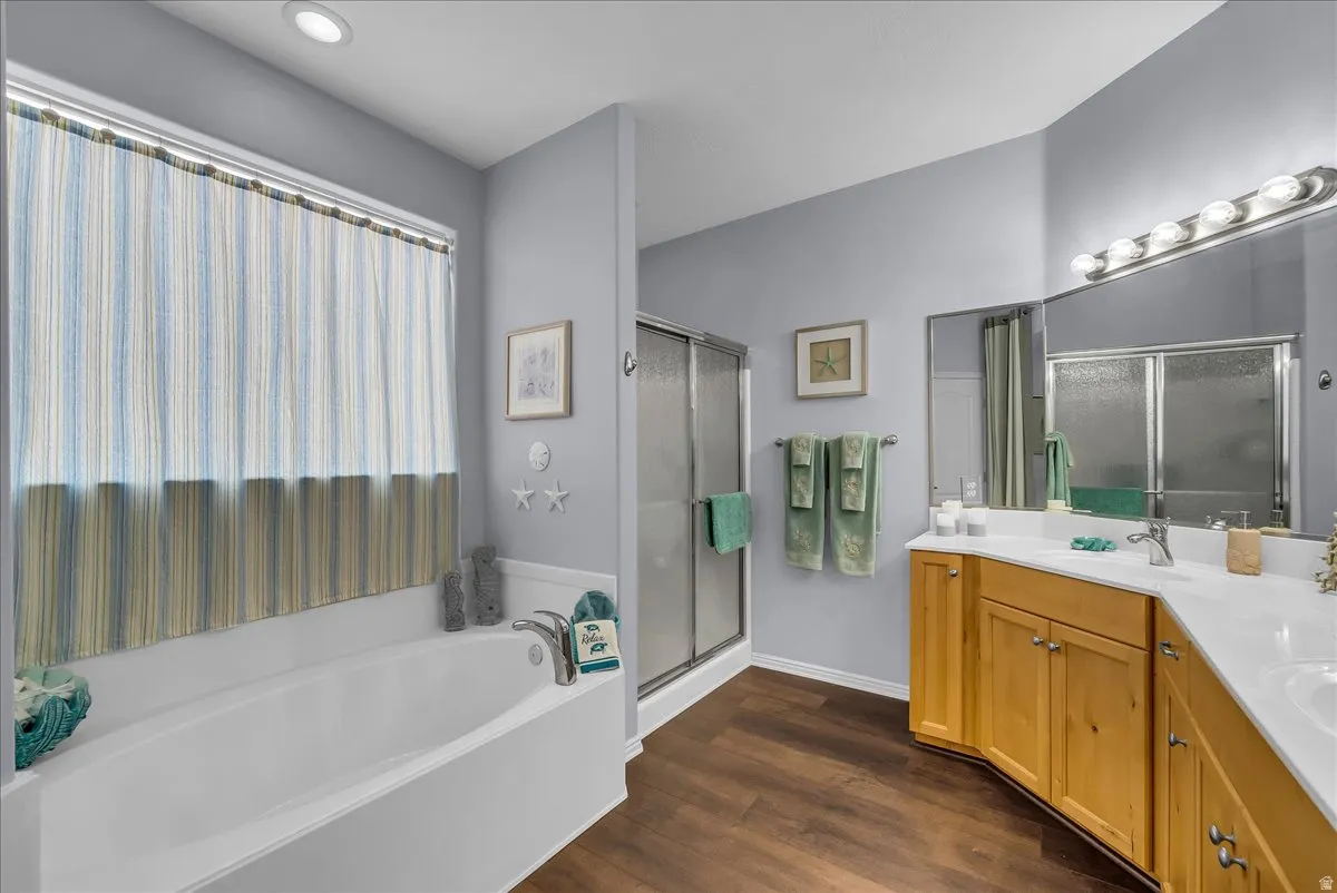 Bathroom featuring a built-in soaking tub, a separate glass-enclosed shower, wood-finish flooring, a double vanity with light wood cabinetry, and a large mirror