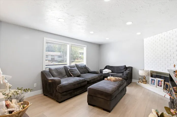 Living area featuring light wood-finish flooring, white brick fireplace, multiple recessed ceiling lights, and dual windows with blinds