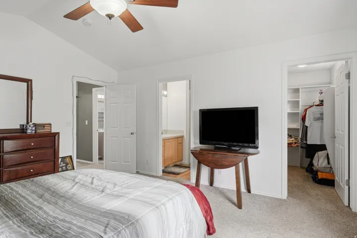 Carpeted bedroom with a ceiling fan, featuring a door opening to a bathroom with a wood-finish vanity and a second door opening to a closet with built-in shelving