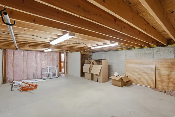 Unfinished basement space featuring exposed wood joists, concrete flooring, fluorescent lighting, and partially insulated walls