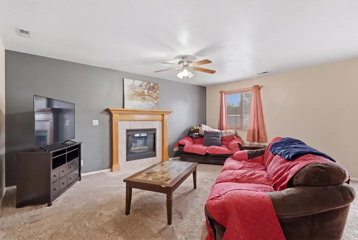 Living area featuring a gas fireplace with a tiled surround and light wood mantel, neutral carpet flooring, and a ceiling fan