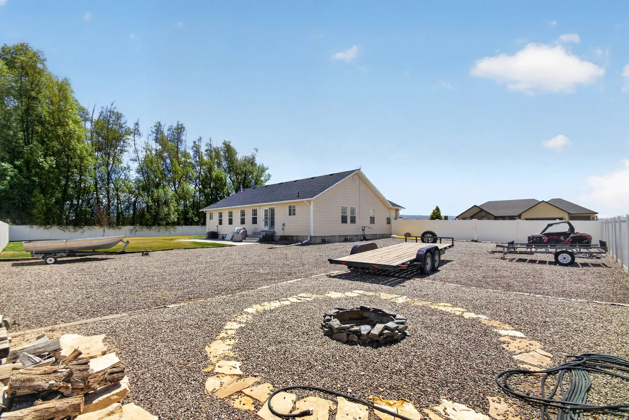 Expansive gravel backyard featuring a stone fire pit, bordered by a white privacy fence and mature trees