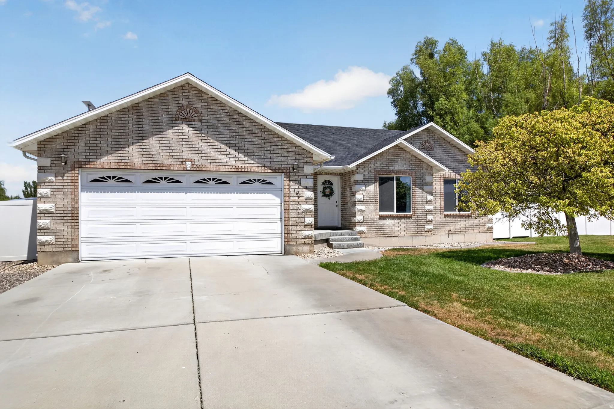 Brick-clad exterior featuring a two-car garage, a concrete driveway, a front lawn with a mature tree, and a white entry door