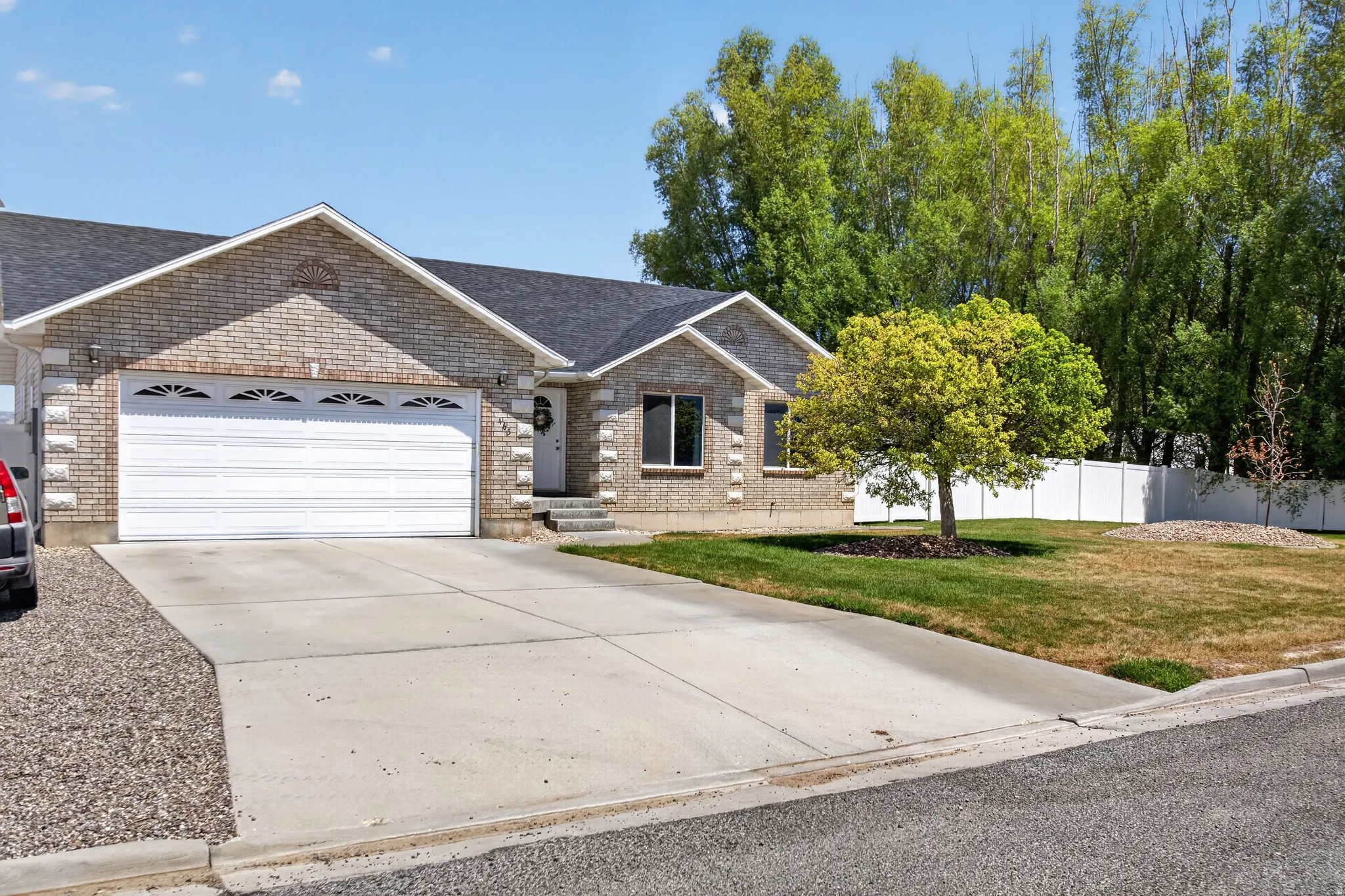 Brick facade residence featuring an attached two-car garage, mature front yard landscaping, and a white privacy fence