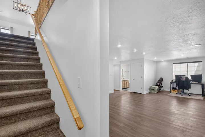 Carpeted staircase with a wood handrail and balusters, opening to a spacious room with wood-finish flooring, recessed lighting, and a large window