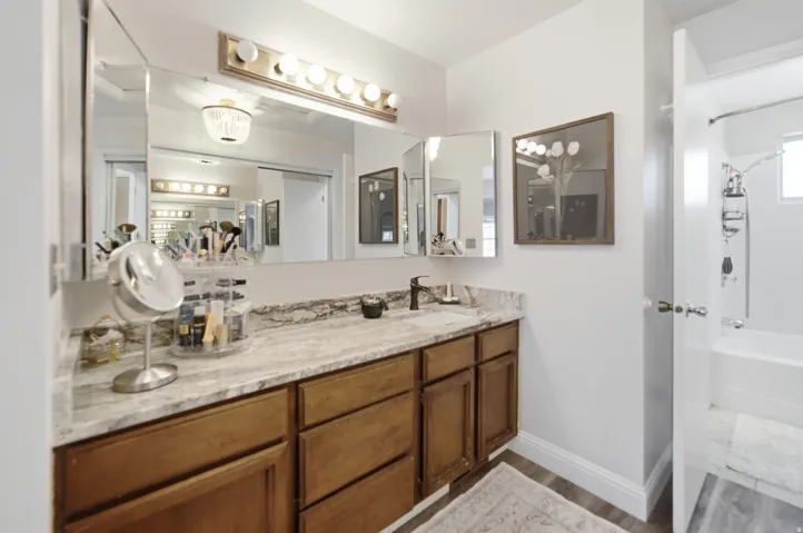 Bathroom vanity featuring a light-toned stone-finish countertop, wood-finish cabinetry, and a large mirror with a multi-bulb vanity light