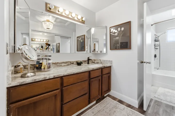 Bathroom vanity featuring a light-toned stone countertop, dark wood-finish cabinetry, and a wide mirror with a strip light fixture
