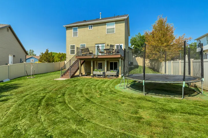 Expansive backyard oasis featuring a manicured lawn, mature landscaping, elevated composite deck with railings, trampoline, and vinyl fencing