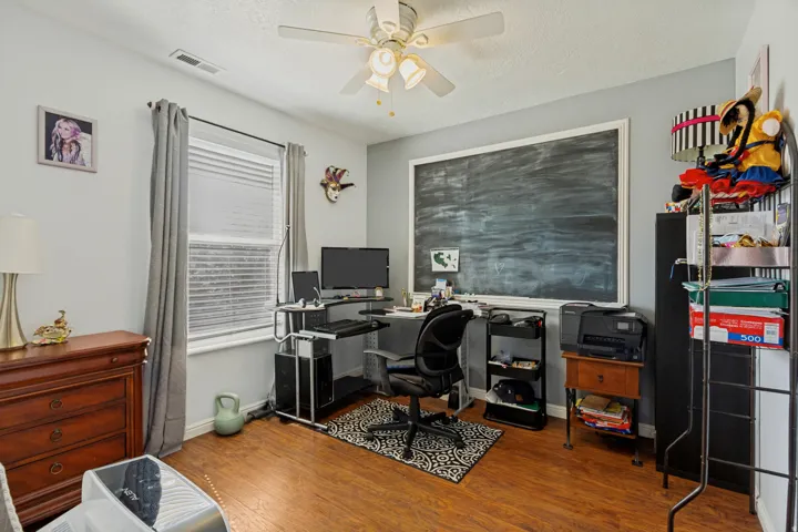 Wood-finish flooring, ceiling fan with light fixture, window with blinds and drapes, and a large framed chalkboard feature