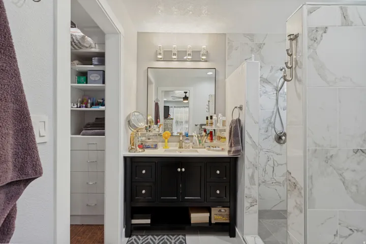 Spa-inspired bathroom featuring a black vanity with multiple drawers and open shelving, white quartz countertop, large framed mirror with integrated lighting, marble-look tile surround, walk-in shower with glass enclosure, and recessed shelving for storag