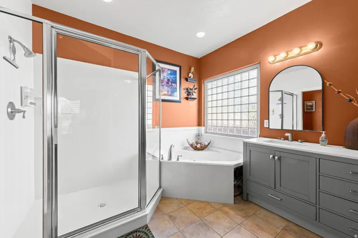 Spacious bathroom featuring a glass-enclosed shower, a large soaking tub, a double vanity with grey cabinetry, tile flooring, and a glass block window