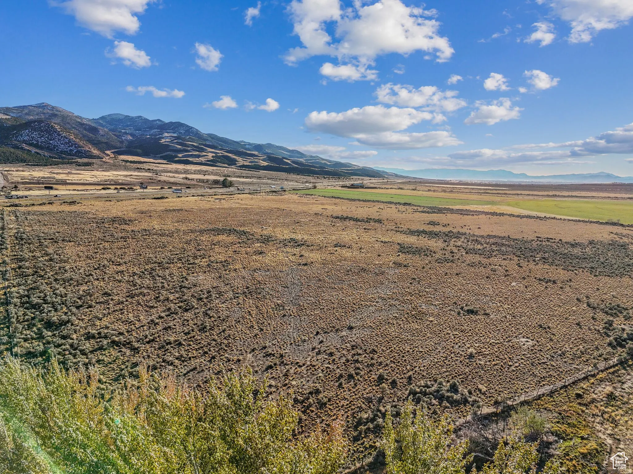 Expansive undeveloped land featuring varied terrain, distant mountains, a partially cultivated field, and a clear blue sky with clouds