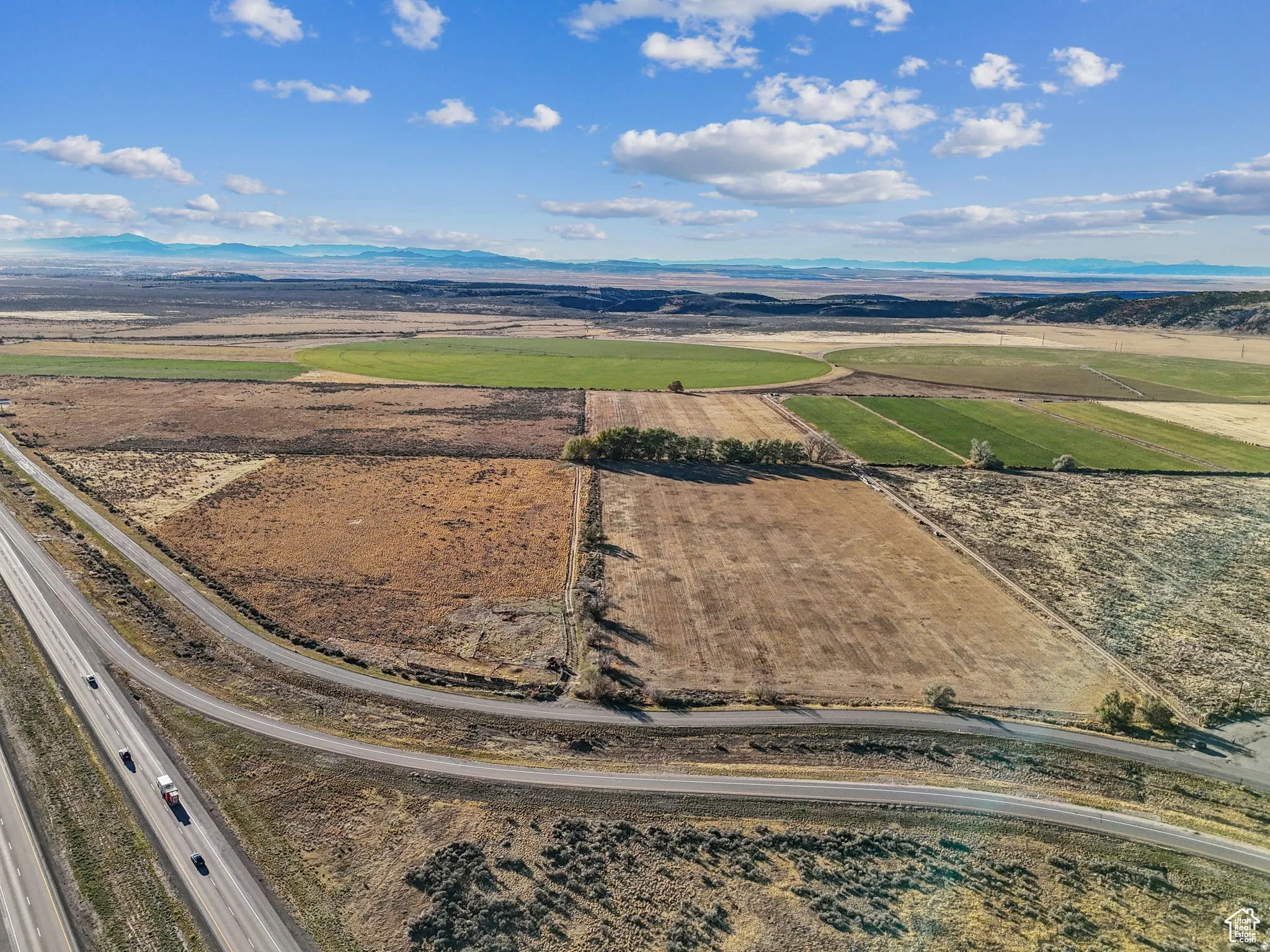 Expansive agricultural land featuring cultivated fields, natural terrain, and distant mountain ranges under a clear sky with scattered clouds