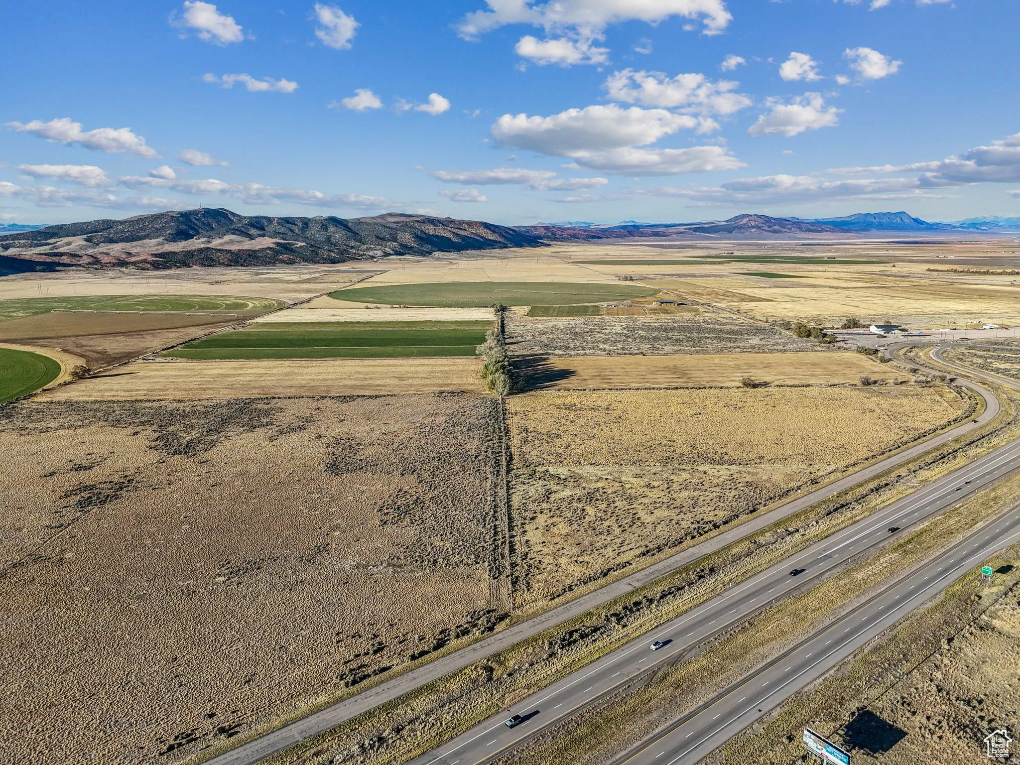Expansive rural landscape featuring a multi-lane highway, agricultural fields with circular irrigation patterns, and distant mountains under a blue sky with scattered clouds