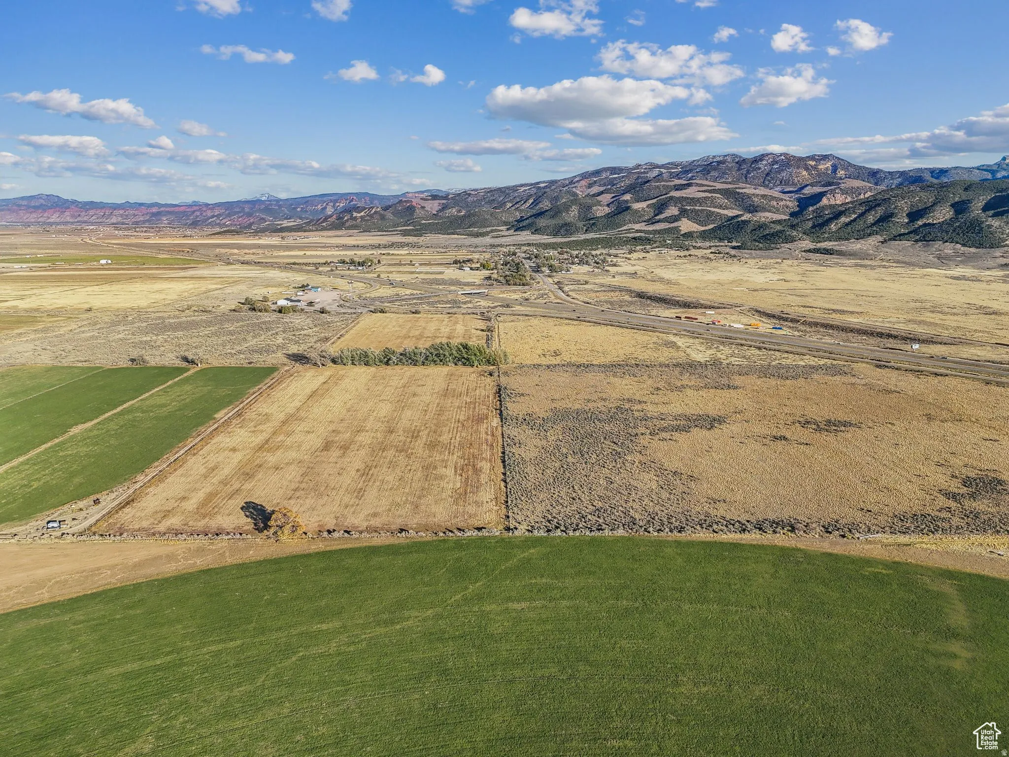 Expansive landscape featuring large agricultural fields, varied terrain, a distant highway, and a mountainous backdrop under a clear blue sky with scattered clouds
