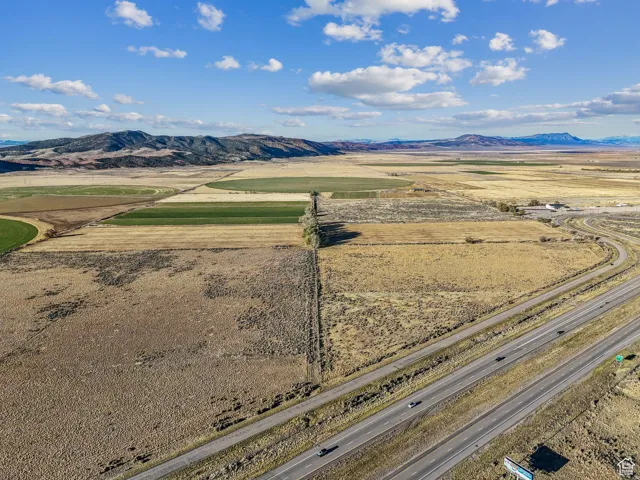 Expansive rural landscape featuring a multi-lane highway, agricultural fields with circular irrigation patterns, and distant mountains under a blue sky with scattered clouds