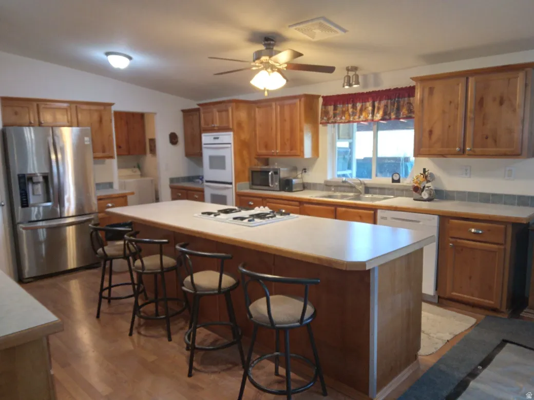 Kitchen featuring wood cabinetry, stainless steel refrigerator, double wall ovens, island with cooktop, and wood-finish flooring