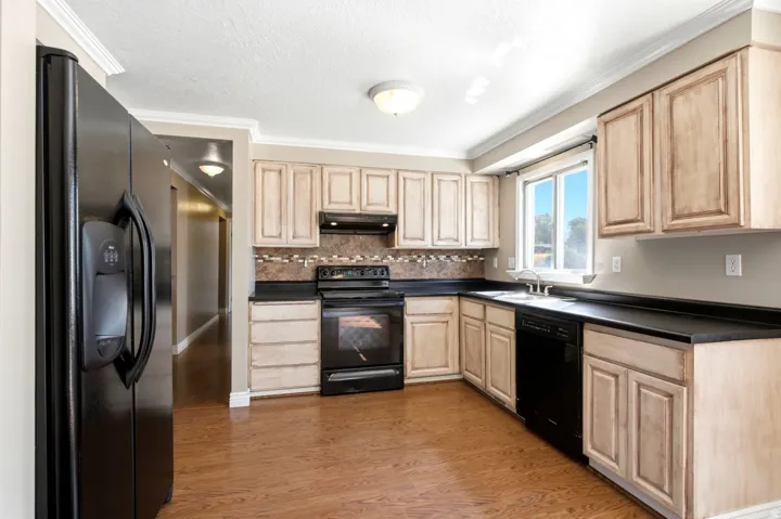 Kitchen featuring wood-finish flooring, light-toned cabinetry, dark countertops, dark appliances, and a window above the sink