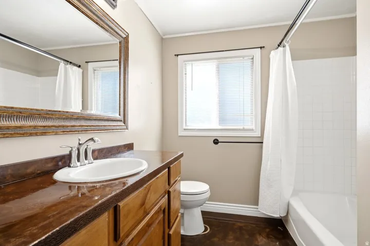 Bathroom featuring a wood-finish vanity with an integrated oval sink and chrome faucet, a large framed mirror, a tub with subway tile surround, a window with blinds, and dark flooring